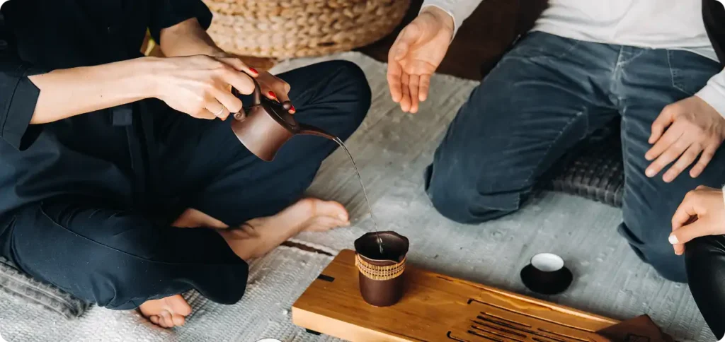 Dwóch mężczyzn siedzi na podłodze, przed nimi naczynia do ceremonii parzenia herbaty, jeden z nich nalewa herbatę.